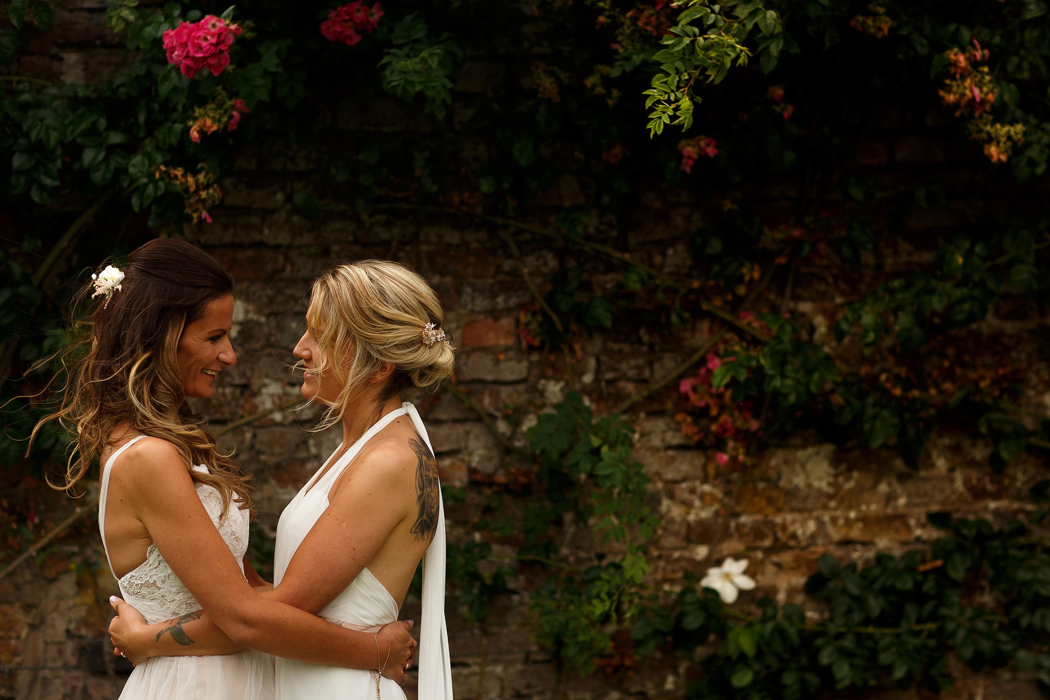 Brides wearing dresses hugging in the gardens at great hall at mains with red brick wall and flowers behind