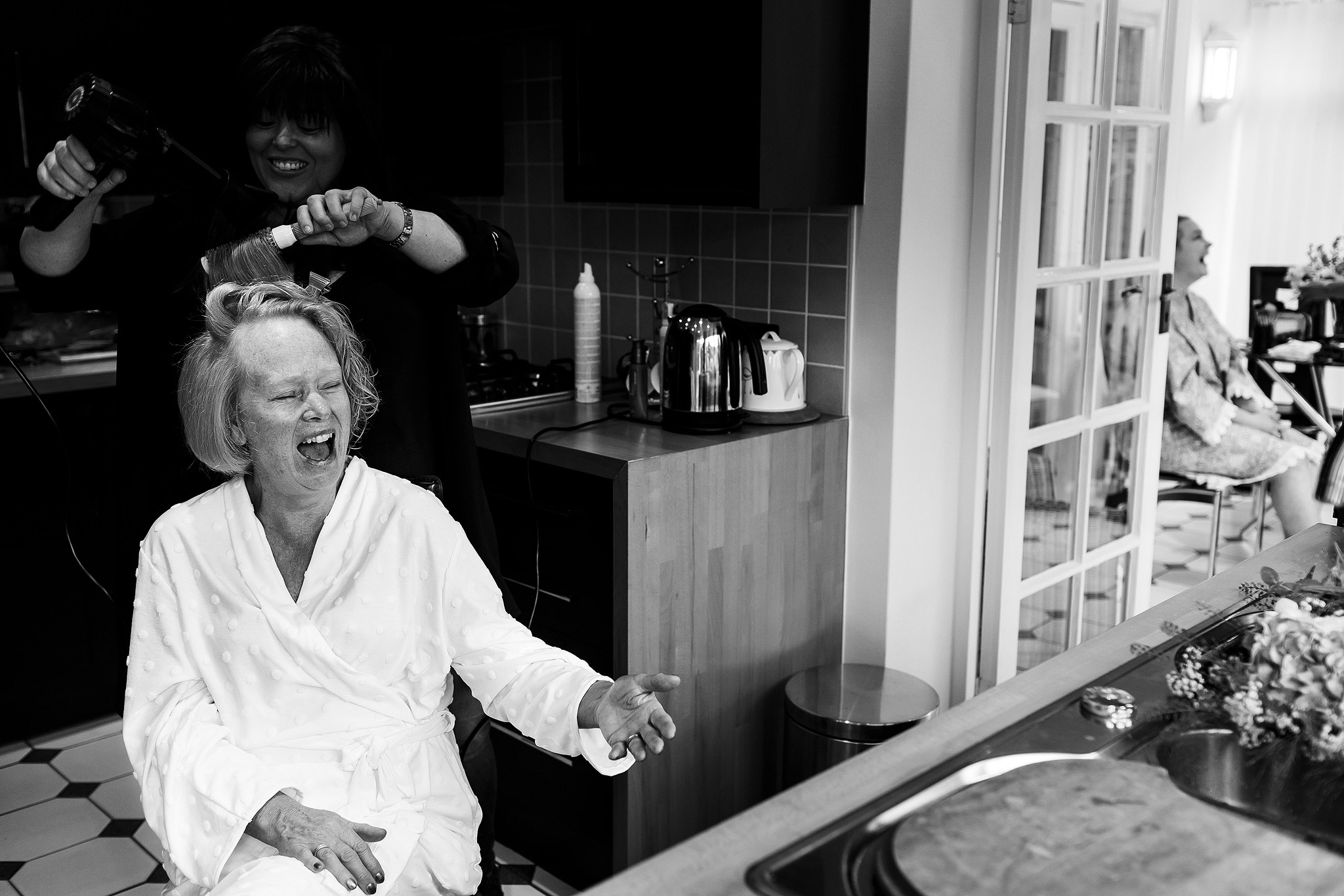 Black and white image of mother of the bride laughing on the morning of her wedding whilst having make up applied