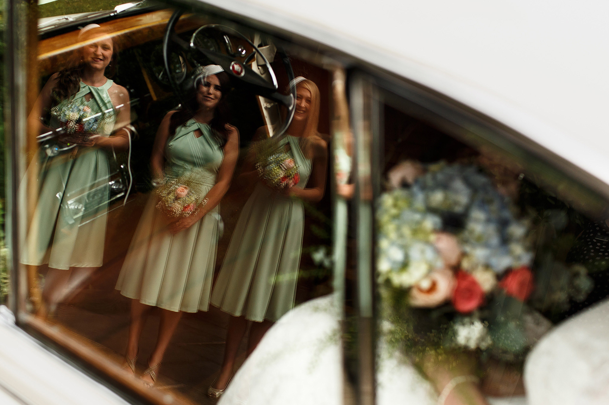 Bridesmaids wearing mint green knee length dresses holding multicoloured wild bouquets waiting for bride to step out of vintage wedding car 