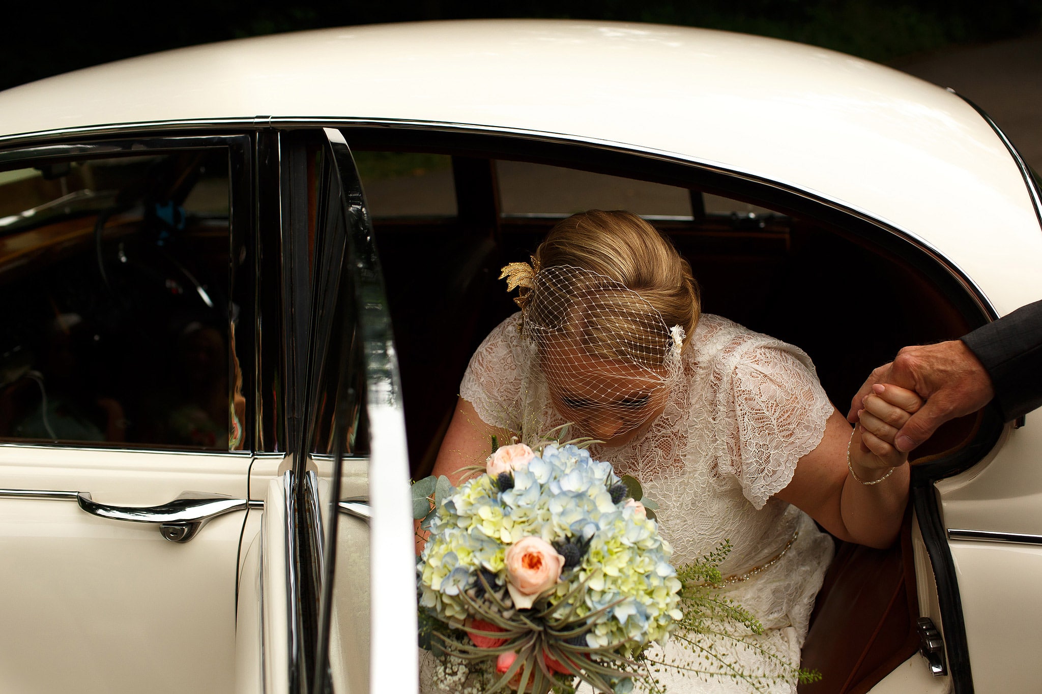 Bride wearing birdcage veil stepping out of vintage wedding car 
