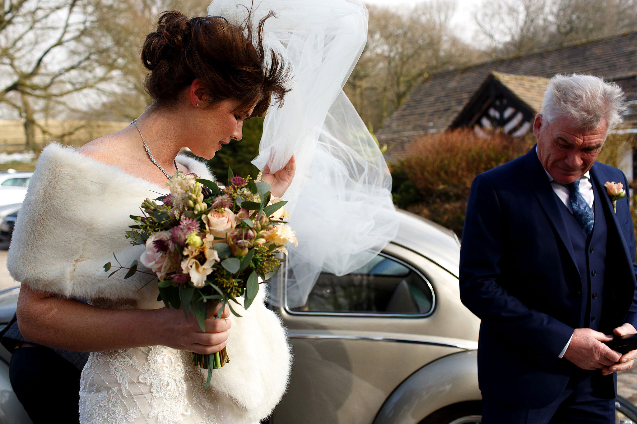 Bride with brunette hair wearing lace strapless wedding dress and fur rug at winter wedding stepping out the wedding car as her long veil blows in the wind.