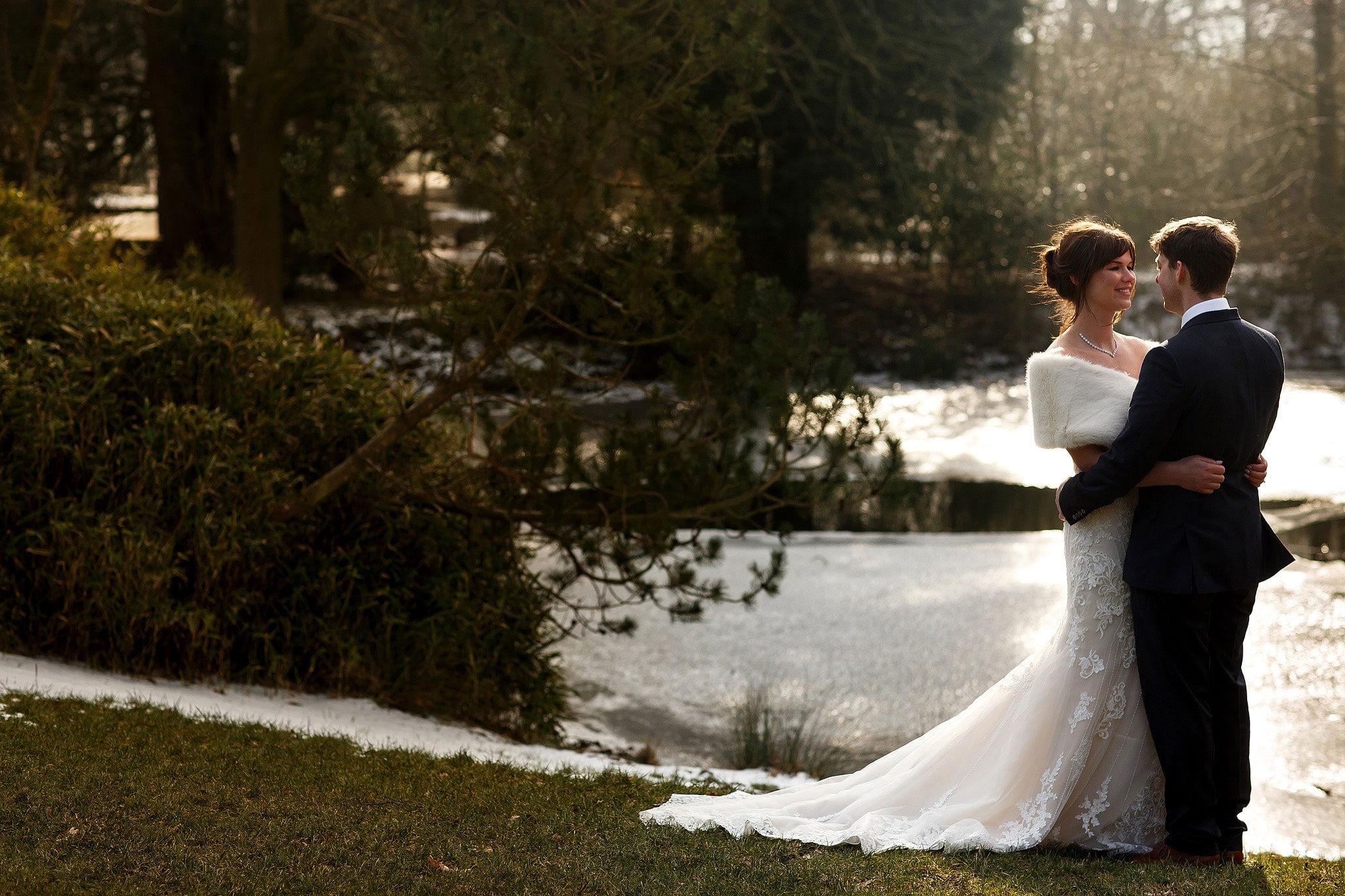 Wedding portrait of bride wearing strapless lace wedding dress and fur shrug in the arms of her husband on a cold winters wedding day with the lake in the background at Rivington Hall Barn
