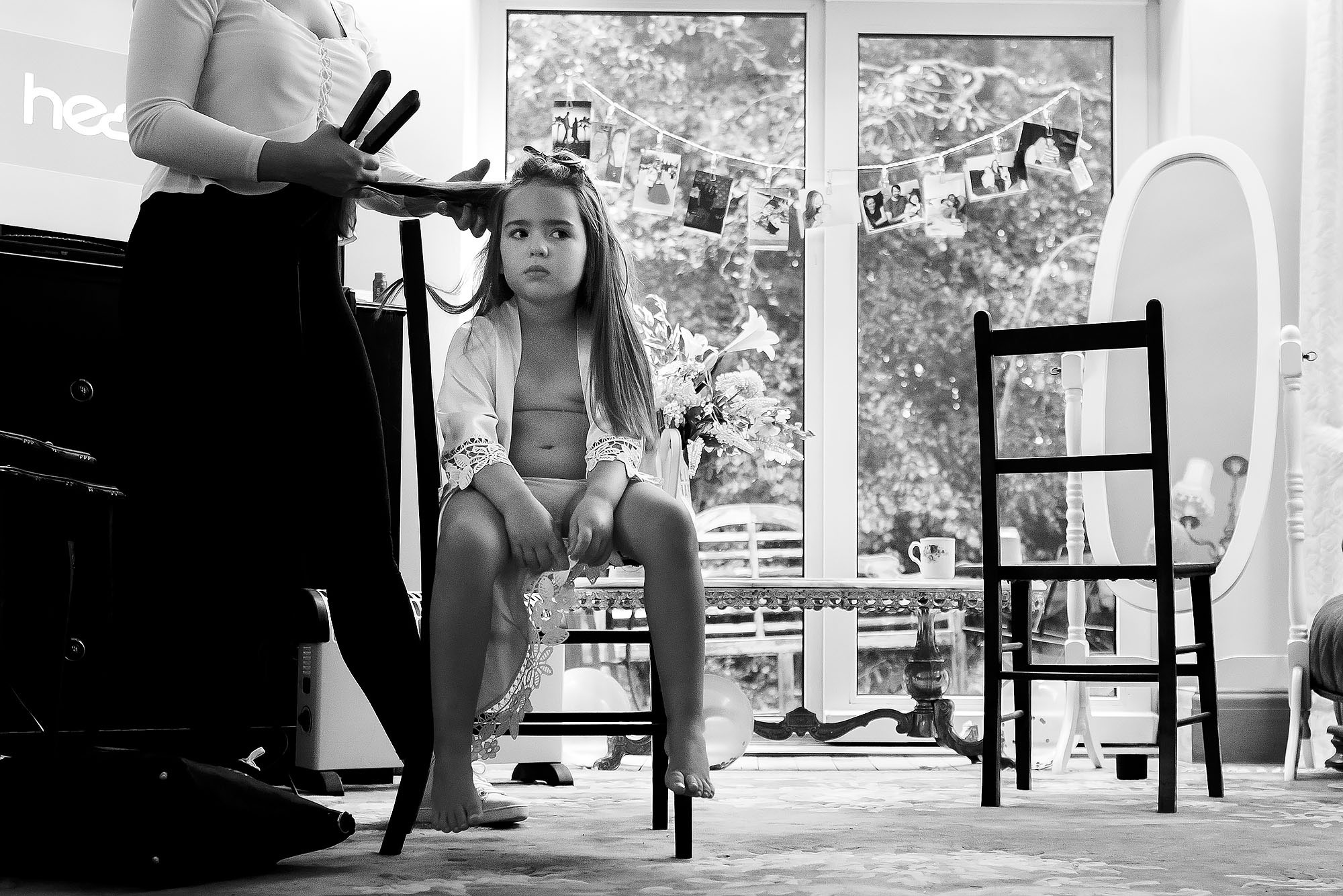 Flower girl pulling grumpy face as she sits legs dangling on a chair having hair done on wedding morning | Wellbeing Farm wedding photographs by Toni Darcy Photography