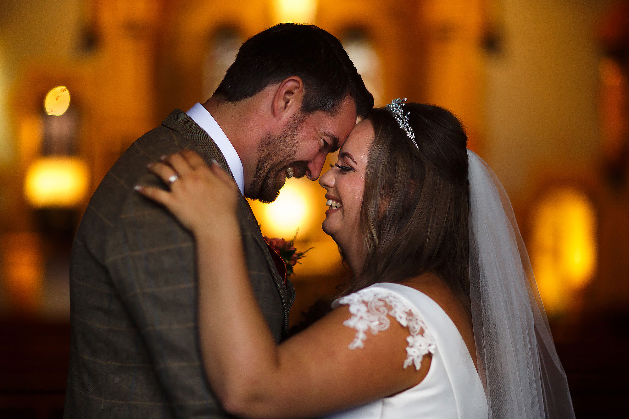 Bride with brunette hair and capped sleeved wedding dress touching forehead with her tall groom inside church with warm lights and candles behind | Wellbeing Farm wedding photographs by Toni Darcy Photography