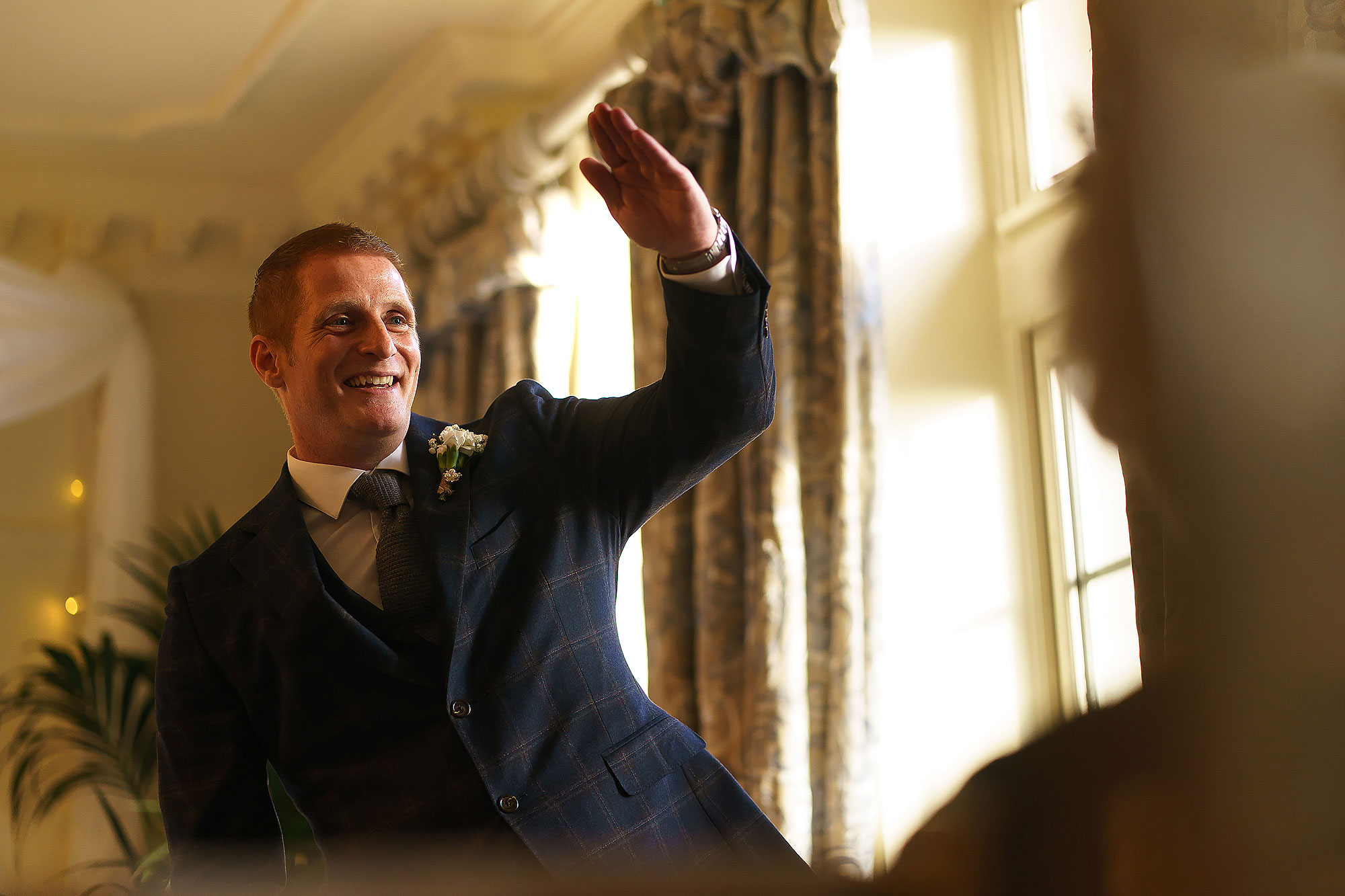 Groom with ginger hair wearing checkered Navy suit waving at guests from the top of the wedding aisle | Eaves Hall wedding photography by Toni Darcy