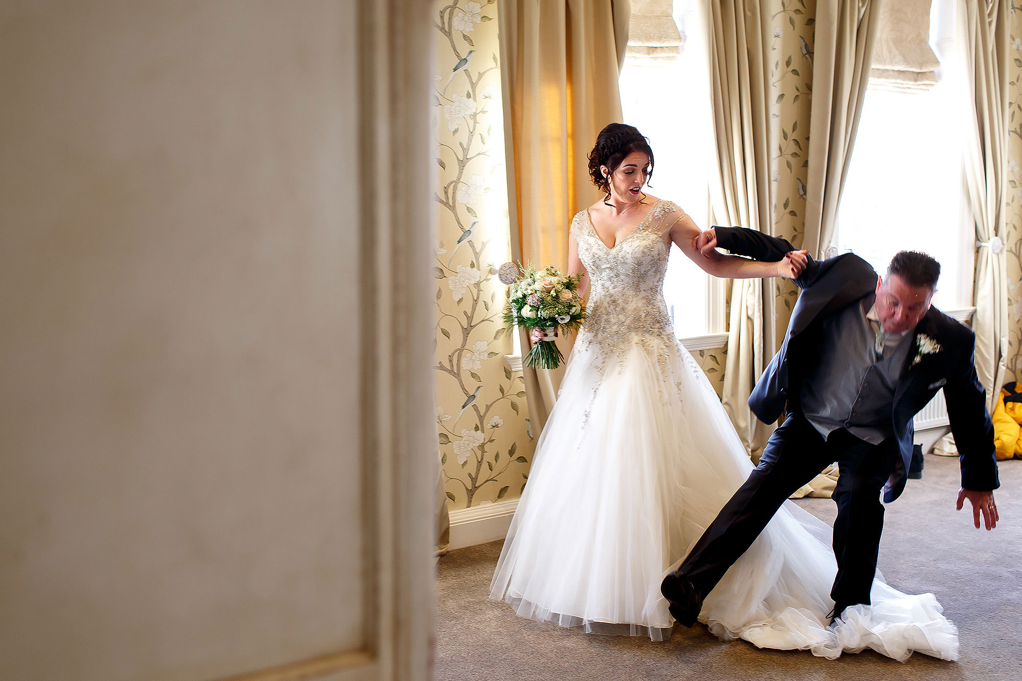 Bride wearing beaded a line wedding dress holding onto the arm of her father as he falls backwards over her wedding dress train | Eaves Hall wedding photography by Toni Darcy