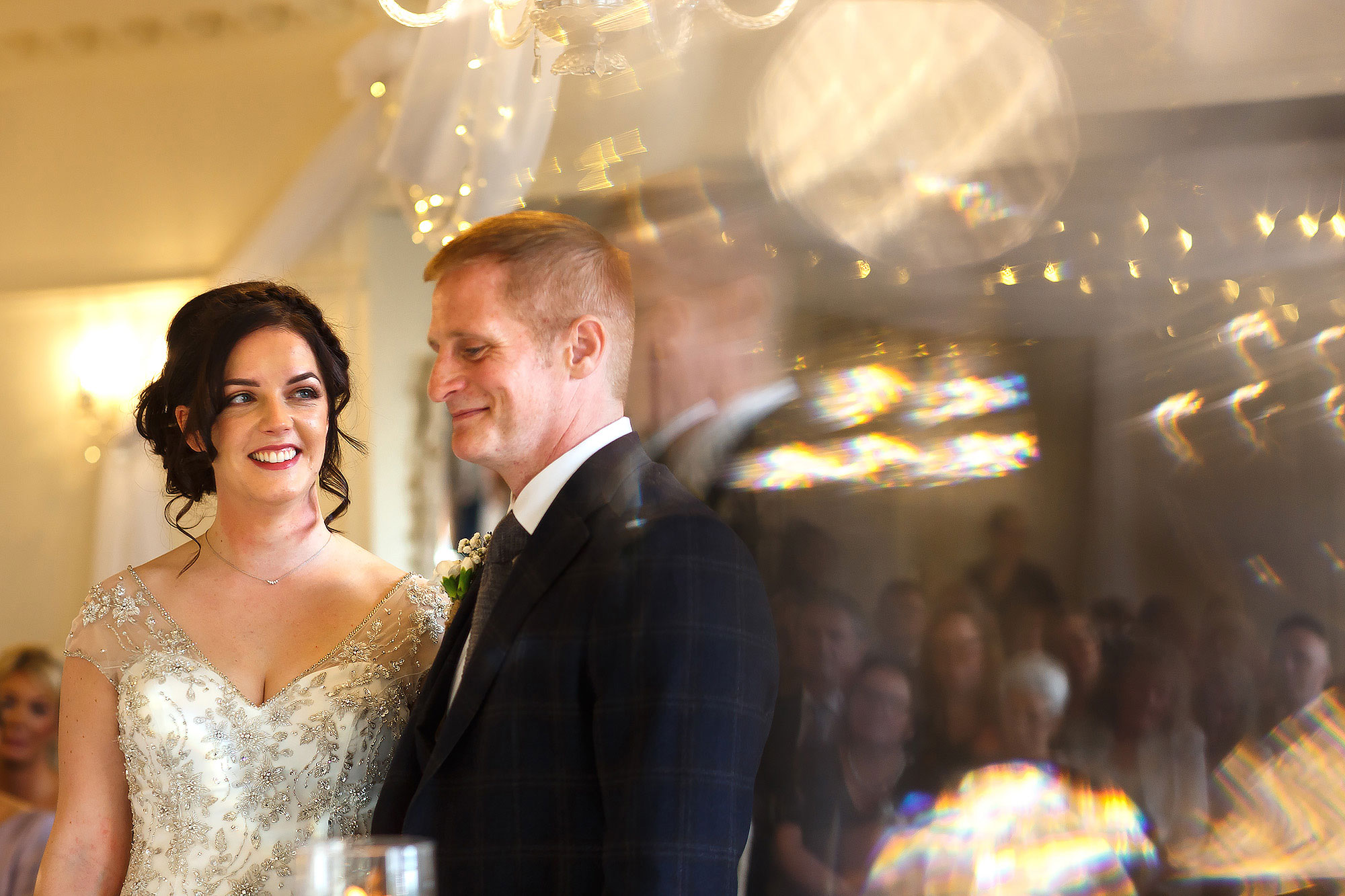 Bride with dark hair and low bun wearing beaded wedding dress smiling, looking towards the groom | Eaves Hall wedding photography by Toni Darcy
