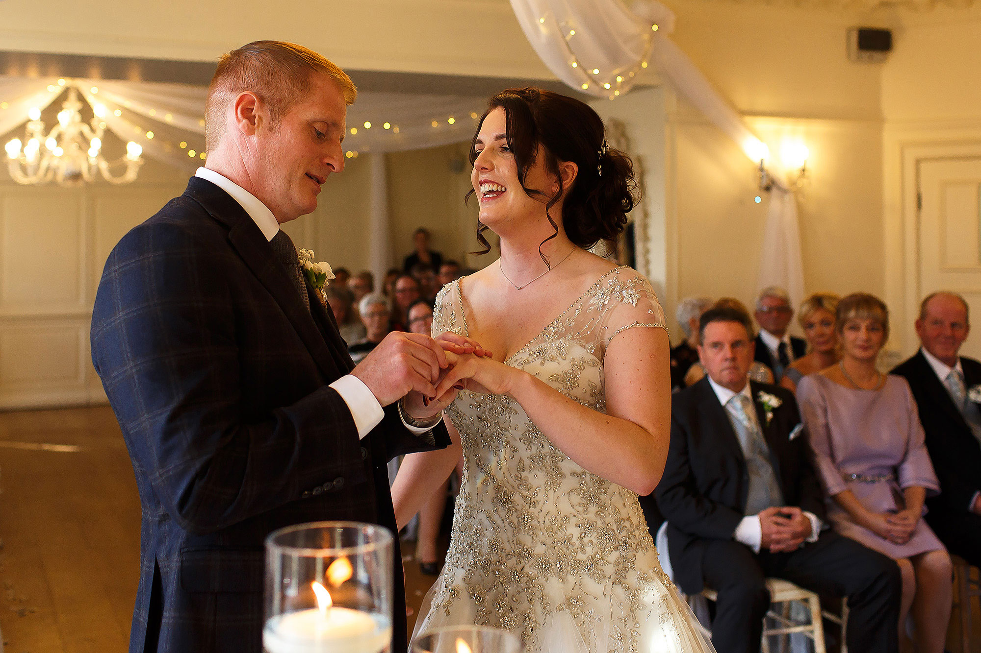 Groom with ginger hair and navy checkered suit placing the ring on brides finger as she laughs wearing beaded wedding dress | Eaves Hall wedding photography by Toni Darcy